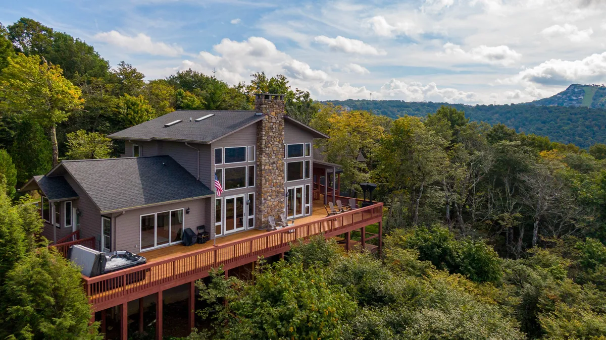 The image showcases the exterior of a spacious mountain home with a large deck overlooking breathtaking panoramic mountain views. Expansive windows invite the outdoors in, while a stone chimney hints at cozy evenings by the fire. The home is nestled among mature trees, with ample space on the deck for gatherings, relaxation, and taking in the serene natural surroundings.