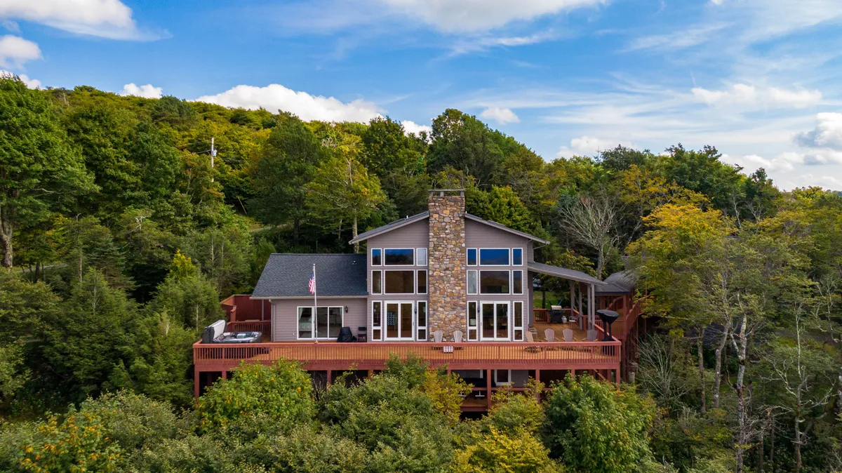 This image features a welcoming wooden deck attached to the chalet, framed by large glass windows and a stone chimney, with lush surrounding trees. The outdoor seating area invites guests to unwind while taking in nature, and the deck provides a perfect vantage point for sunset or stargazing. The American flag adds a touch of warmth and hospitality to the scene.