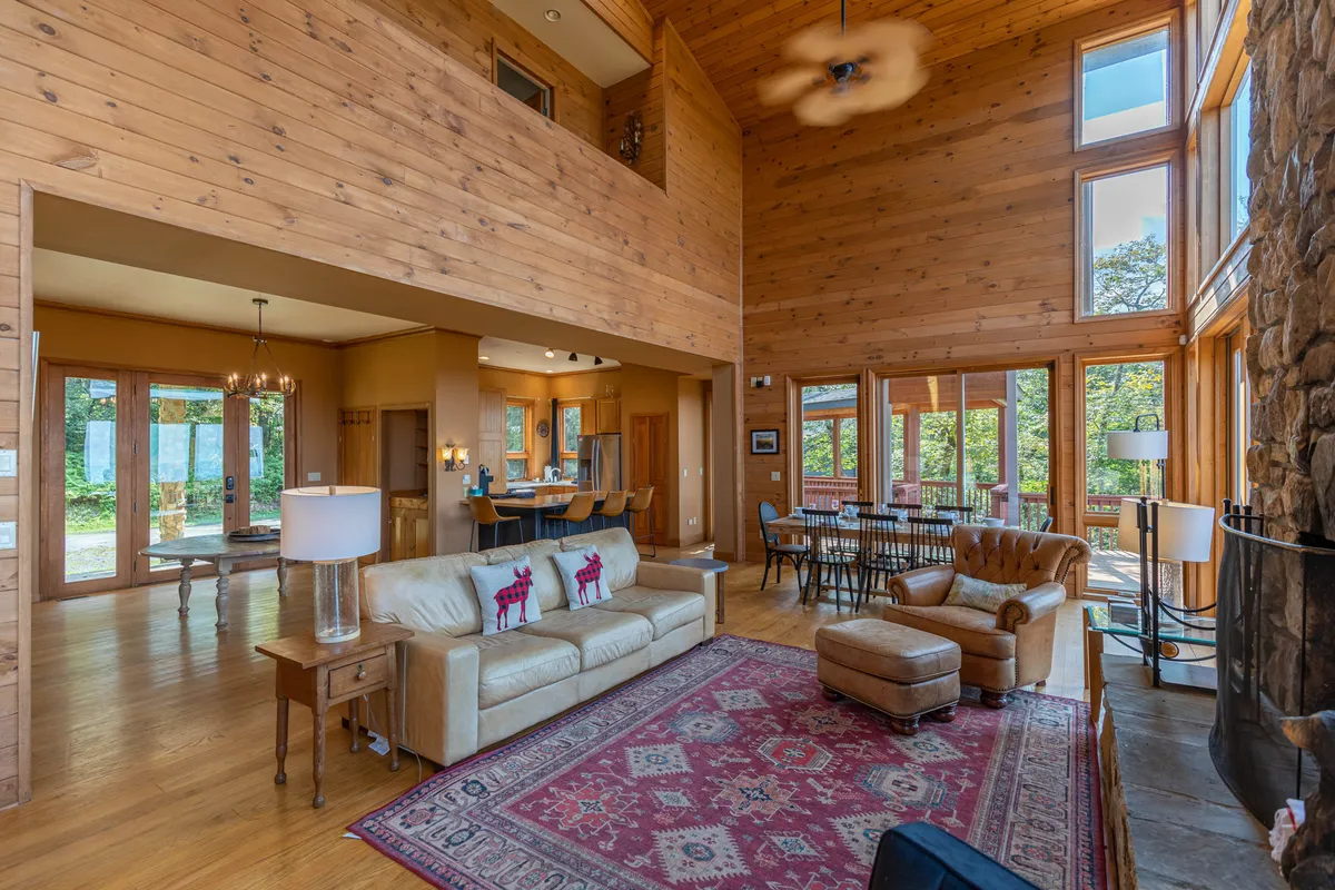 A welcoming living area with a stone fireplace, broad windows offering natural light, a comfortable sofa with decorative pillows, and a dining area in the background. The setting is both elegant and homey, suitable for relaxing or entertaining.