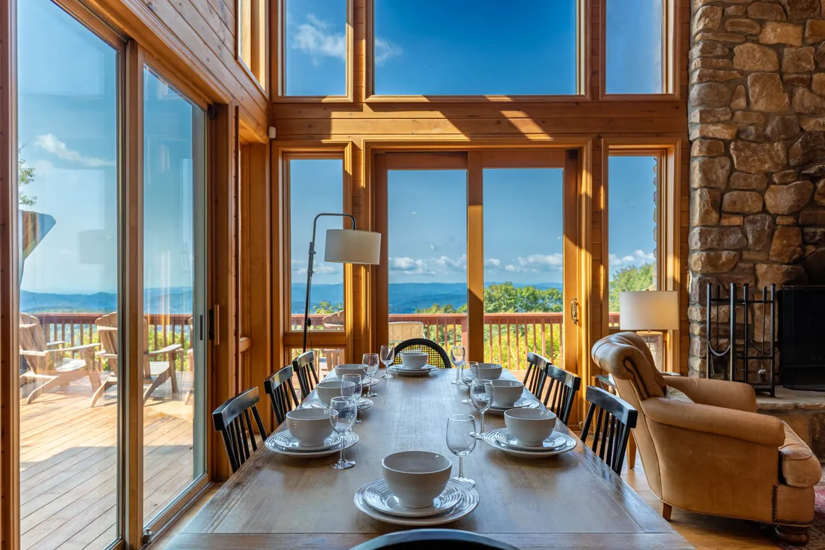 An elegant dining area with a large wooden table set beneath floor-to-ceiling windows that offer sweeping views of the mountains. The stone fireplace in the background and the relaxed seating signal a space designed for meals, conversation, and togetherness.