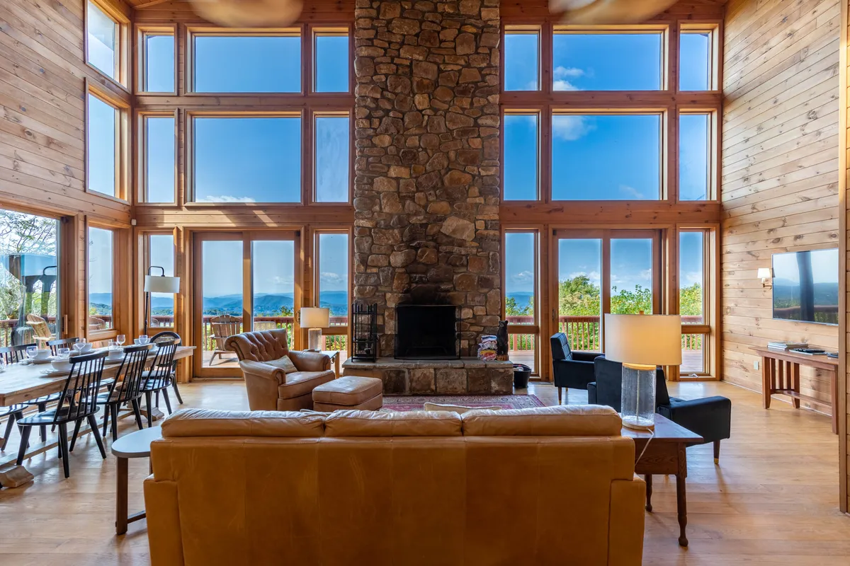 A spacious great room featuring a large stone fireplace, floor-to-ceiling windows that reveal breathtaking mountain views, a wooden dining table, and a comfortable seating area with plush furnishings. Warm natural light pours in, highlighting the open layout and inviting ambiance perfect for gatherings.