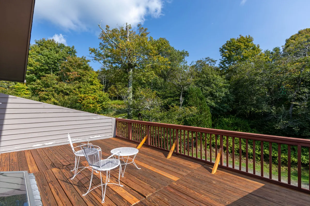 A wooden deck furnished with white metal chairs and a round table, set against a lush greenery backdrop. The inviting space is ideal for morning coffee, reading, or sunset gatherings, making it a versatile outdoor retreat.