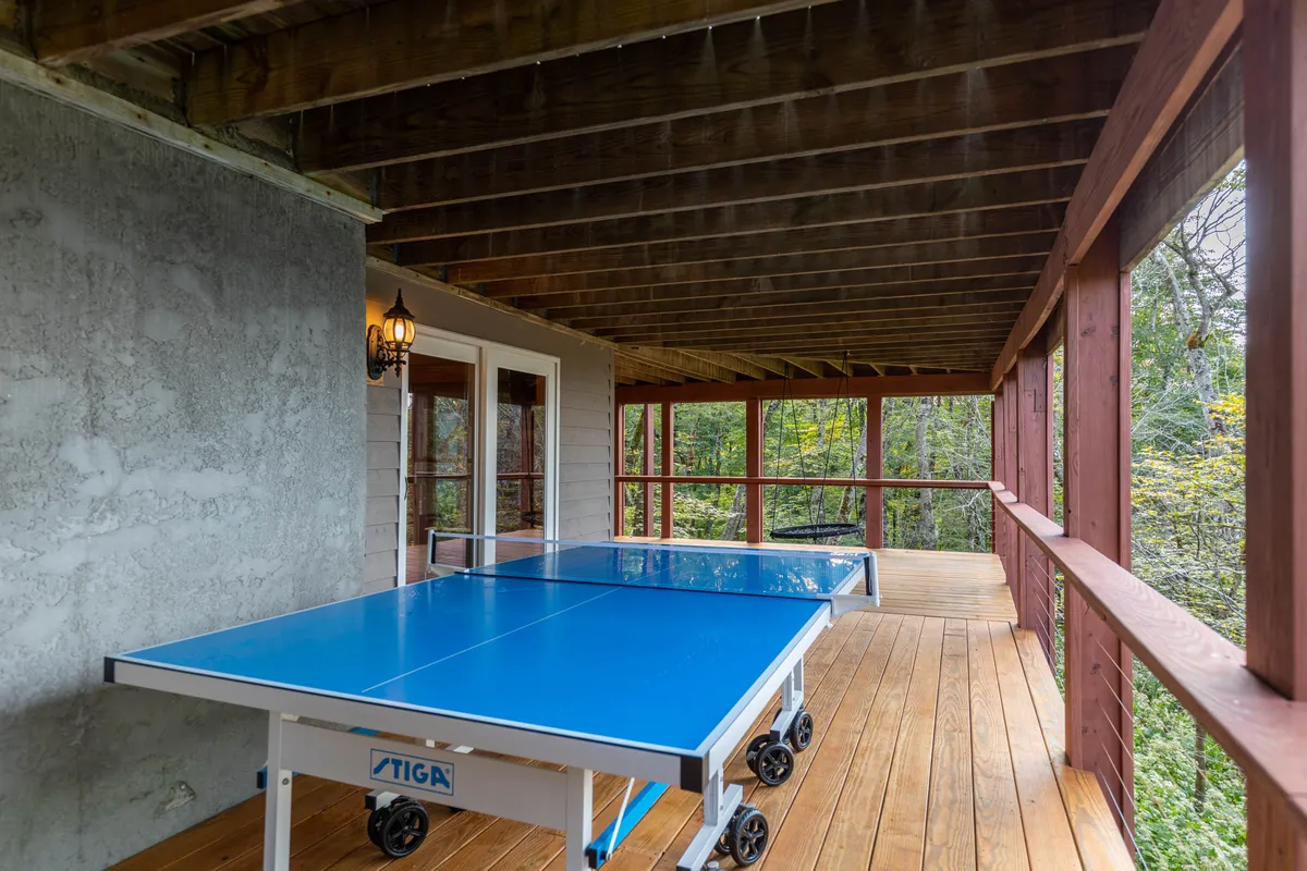A playful exterior scene featuring a ping pong table on a wooden deck, surrounded by trees and dappled light. The relaxed setup encourages friendly competitions and outdoor fun, ideal for families and groups with children.