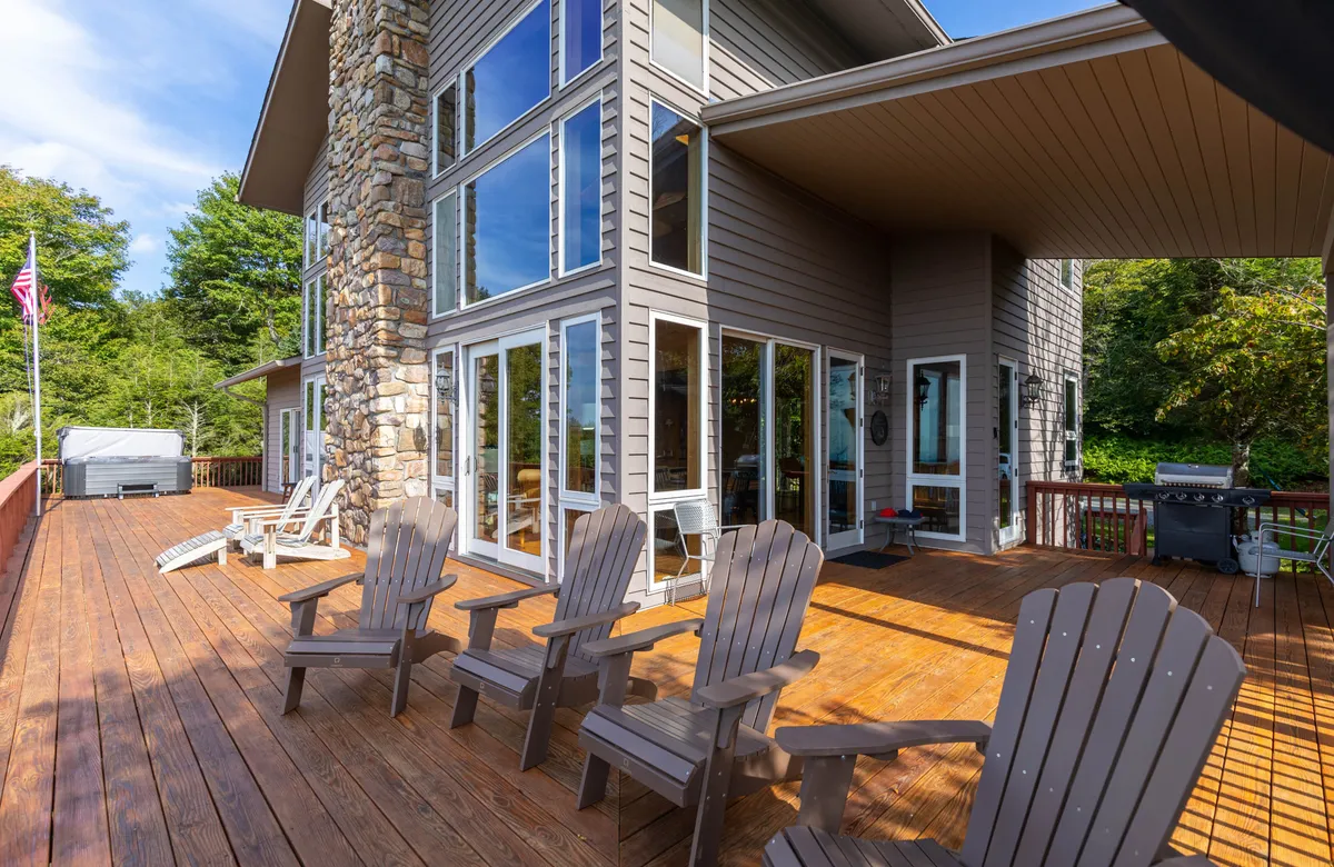 This exterior shot highlights the home's expansive wooden deck with Adirondack chairs, large glass windows, and a stone accent wall. The setting is surrounded by lush trees, projecting a tranquil mountain retreat ambiance. The open deck invites gatherings and relaxation, while the impressive windows hint at panoramic interior views.